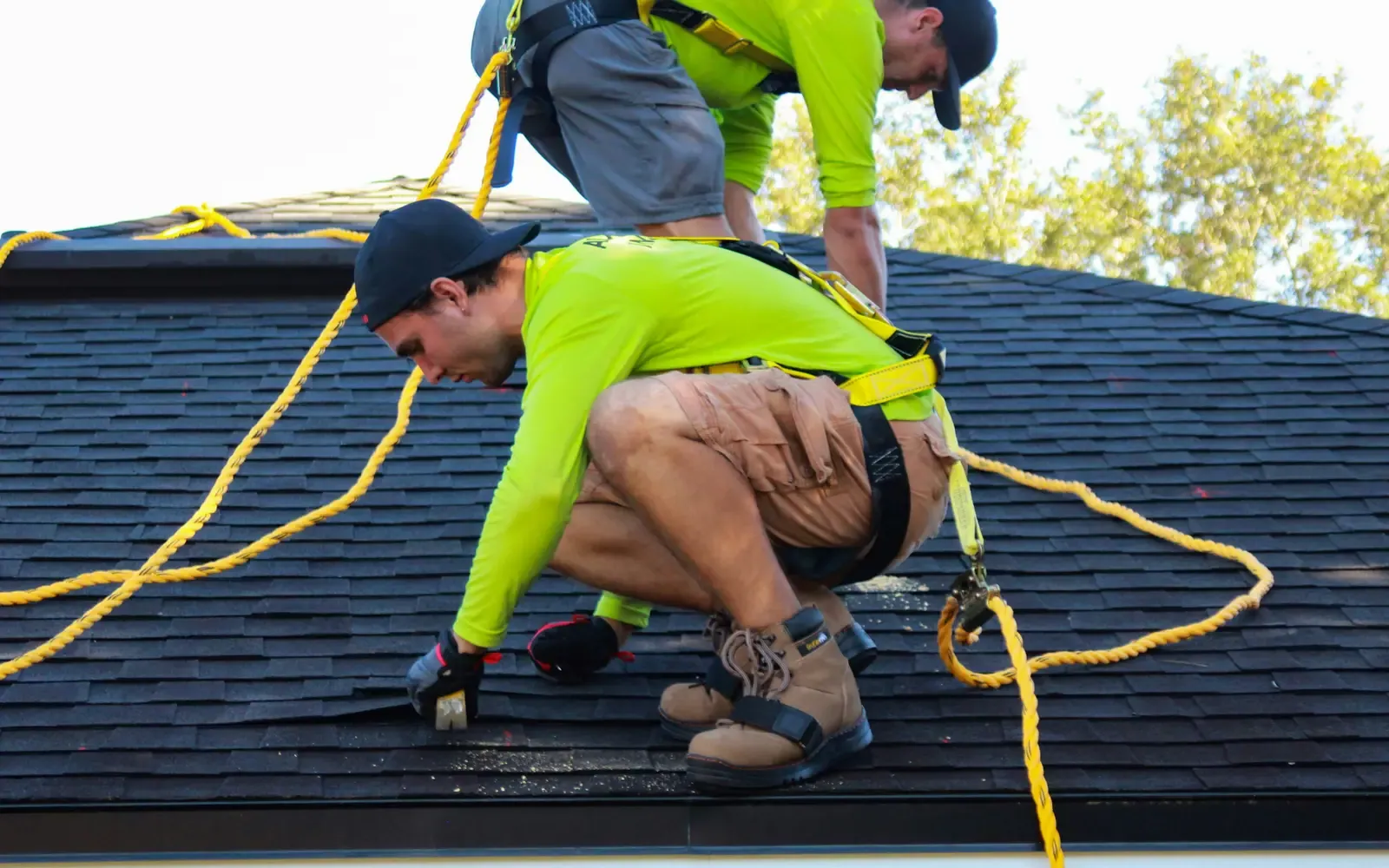 Two roofers in safety harnesses installing asphalt shingles on a house roof
